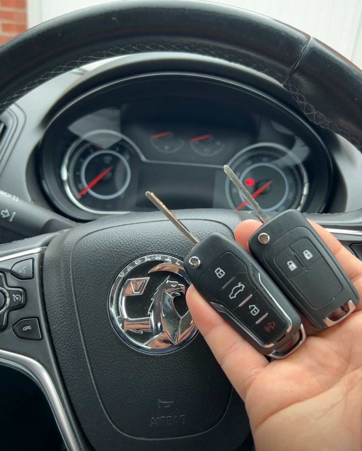 Person holding two car keys in front of a steering wheel with a Vauxhall emblem.